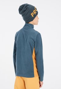 Fleece pullover i teal med en mock neck, der har et gult sidepanel. En matchende teal beanie har "PRO" trykt i fed gult.
