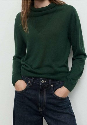 Kvinde iført en mørkegrøn langærmet sweater, stoppet ned i højtaljede mørkeblå jeans med hænderne i lommerne.
