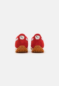 Red suede athletic shoes with white accents and a golden logo. Textured rubber outsole with circular protrusions. Rear view.