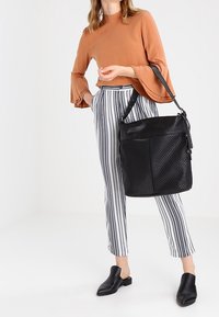 Zwarte geperforeerde tote tas met een handvat bovenaan en een verstelbare schouderband. Draag het met gestreepte broek en een lange mouw roestkleurige top.