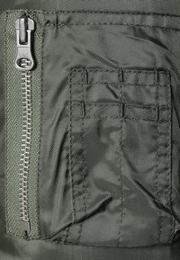 Tissu vert olive à finition texturée, doté d'une fermeture éclair centrale et d'une poche cousue avec des lignes horizontales. Design durable et fonctionnel.