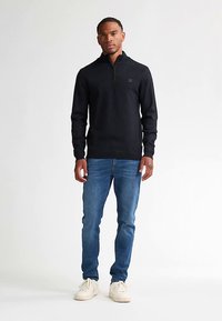 Maglione nero con zip in tessuto liscio, abbinato a jeans di denim blu. Scarpe da ginnastica bianche completano l'outfit casual.