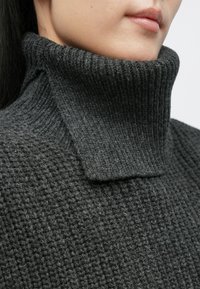 Donkergrijze gebreide trui met een hoge, geribbelde kraag en een gestructureerd oppervlak. Opvallend vanwege de dikke, warme stof en het gestructureerde ontwerp.