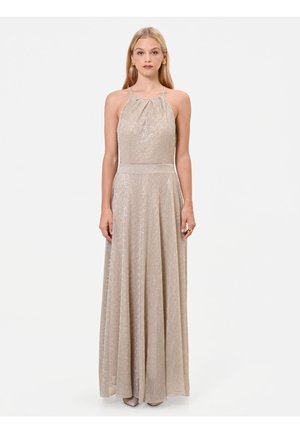 Donna con un abito beige metallizzato senza maniche, lungo fino a terra, con scollo halter, in piedi su uno sfondo bianco semplice.
