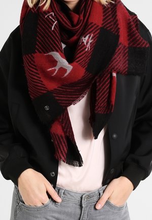 Écharpe écossaise rouge et noire avec motifs de cerfs blancs et motifs géométriques portée sur une veste noire et une chemise rose clair, mains dans les poches d'un jean gris.