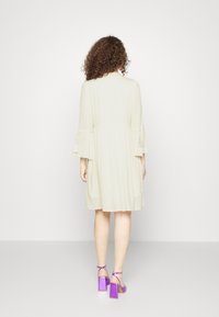 Ljust beige klänning med plisserad textur och trekvartslånga ärmar, med knyt detaljer vid ärmsluten. Matchad med lila klacksandaler med blockklack.