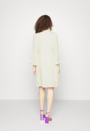 Hellbeiges Kleid mit plissierter Textur und Dreiviertelärmel, mit Bindebändern an den Manschetten. Kombiniert mit lila Blockabsatzsandalen.