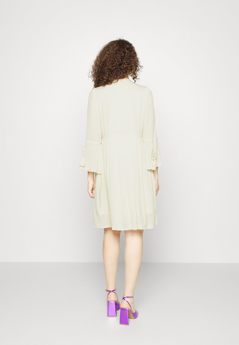 Ljust beige klänning med plisserad textur och trekvartslånga ärmar, med knyt detaljer vid ärmsluten. Matchad med lila klacksandaler med blockklack.