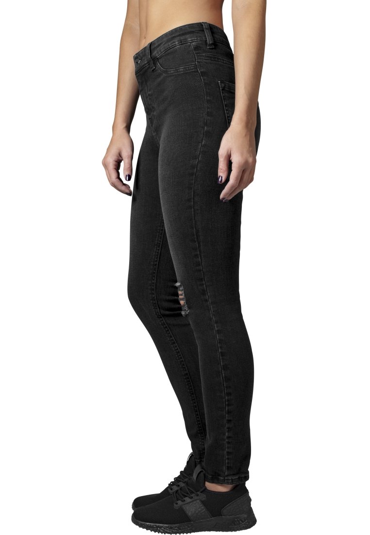 Fekete, skinny farmernadrág magas derekú fazonnal, amelyen kopott részletek találhatók a combokon, denim anyagból készült. Fekete sportcipőkkel kombinálva.
