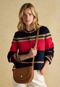 Femme aux cheveux blonds portant un pull à rayures bleu marine, rouge, vert et beige, tenant un sac en cuir marron en bandoulière sur un pantalon blanc.