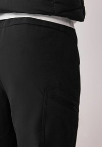 Black cargo pants featuring a smooth texture, a fitted design, and a zippered pocket in an angled shape on the thigh.