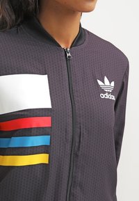 Giacca nera con zip caratterizzata da un motivo testurizzato, con strisce orizzontali bianche, rosse, blu e gialle sul lato sinistro, con il logo Adidas.