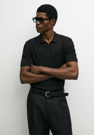 Mann trägt schwarzes kurzärmeliges Strickpolo, schwarzen gewebten Gürtel, dunkle Hose und große schwarze Sonnenbrille, die Arme verschränkt und schaut zur Seite.