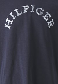 Marineblå bomulls-T-skjorte med et slitt hvitt trykk av "HILFIGER" i en buet form over brystet. Myk tekstur, uformell design.