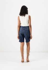 Bermudas de denim con un corte holgado, en color azul oscuro, con dobladillos rematados y dos bolsillos traseros. Se llevan con una blusa sin mangas blanca.