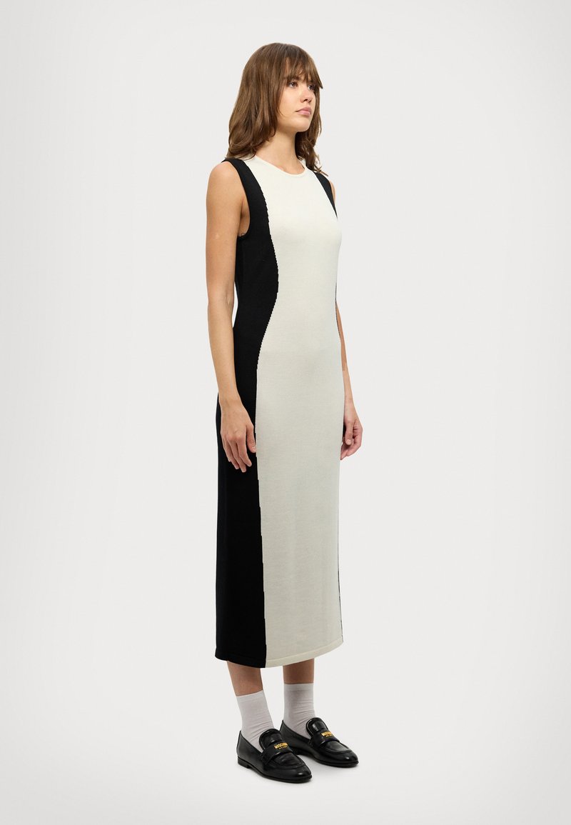 Abito senza maniche, di lunghezza al ginocchio, con un design a blocchi di colore in bianco sporco e nero, in tessuto a maglia con una texture liscia; abbinato a mocassini neri.
