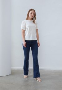 White top with eyelet puff sleeves, paired with dark blue flared jeans and white block-heeled sandals, set against a light background.