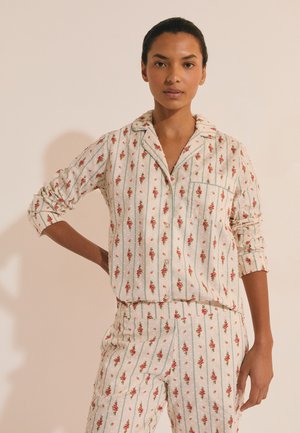 Frau mit dunklem Haar, die ein cremefarbenes, gestreiftes Schlafanzug-Set mit Blumenmuster und hochgekrempelten Ärmeln trägt und vor einem schlichten beigen Hintergrund steht.