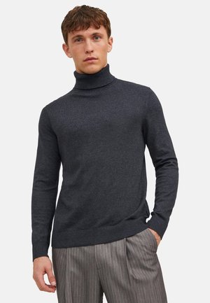 Młody mężczyzna ubrany w ciemnoszary sweter z golfem i szare spodnie w prążki, stojący z jedną ręką w kieszeni na jednolitym tle.