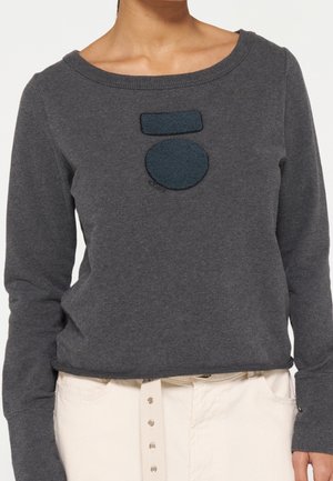 Personne portant un pull à manches longues gris foncé avec un patch circulaire et rectangulaire sur la poitrine, associé à un pantalon beige clair et une ceinture blanche.