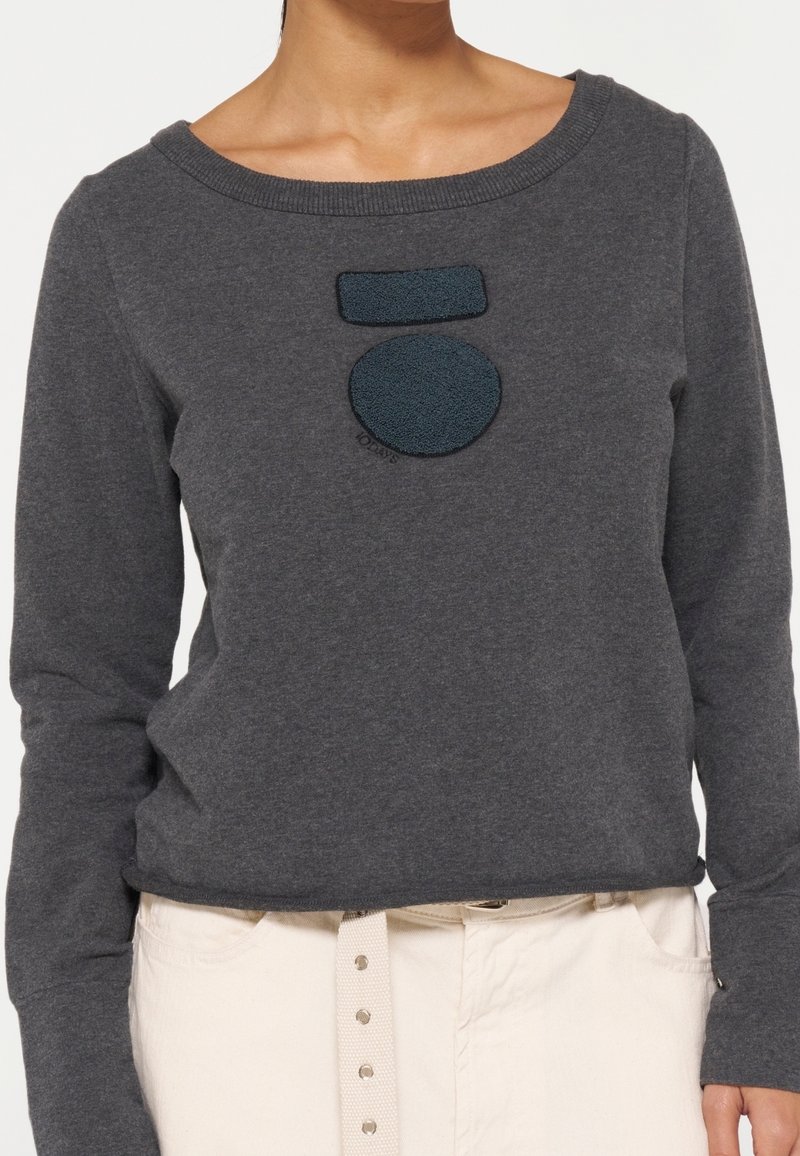Personne portant un pull à manches longues gris foncé avec un patch circulaire et rectangulaire sur la poitrine, associé à un pantalon beige clair et une ceinture blanche.