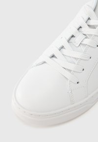 Λευκό αθλητικό παπούτσι με λείο επάνω μέρος από δέρμα, στρογγυλεμένη μύτη, υφασμένο καουτσούκ στη σόλα και επίπεδα κορδόνια από βαμβάκι, με ραφές διακοσμητικές κατά μήκος των άκρων.