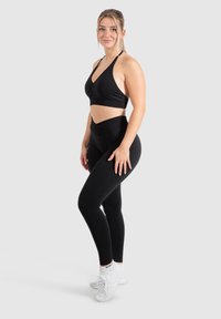 Zwarte sportbeha met een V-hals en gathered detail, gecombineerd met hoge zwarte leggings. Model draagt witte sportschoenen.