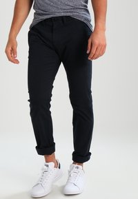 Smalpassande svarta chinos med uppvikta cuffs, i kombination med vita sneakers. Materialet verkar slätt med minimal detaljering och en klassisk femfickorsdesign.
