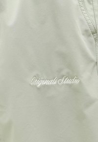 Ανοιχτό μπεζ ύφασμα με λευκό κεντημένο κείμενο «Originals Studio» κοντά στην ραμμένη άκρη της ραφής.