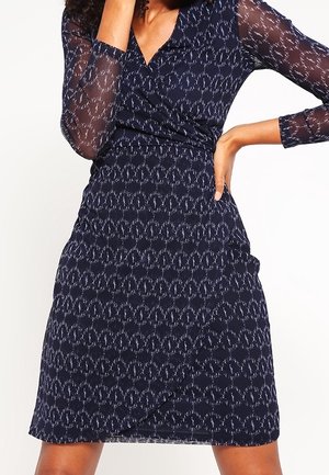 Kobieta w ciemnoniebieskiej sukience kopertowej z przezroczystymi długimi rękawami i subtelnym białym abstrakcyjnym wzorem, pozująca z jedną ręką na biodrze.