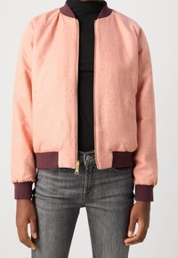 Roze bomberjack met een textuur, geribbelde wijnrode manchetten en kraag, en een ritssluiting aan de voorkant. Draagt over een zwarte top, gecombineerd met grijze jeans.