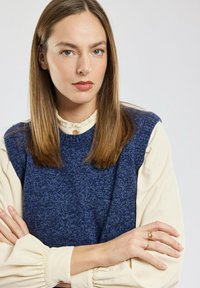 Granatowy sweter bez rękawów z dzianiny nałożony na kremową bluzkę z długim rękawem z puszystymi rękawami i falbanowym kołnierzykiem; złoty pierścionek na ręce modelki.