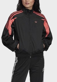 Svart zip-up-jacka med rosa detaljer och tre vita ränder på ärmarna. Har en hög krage och elastiska muddar och nederkant.