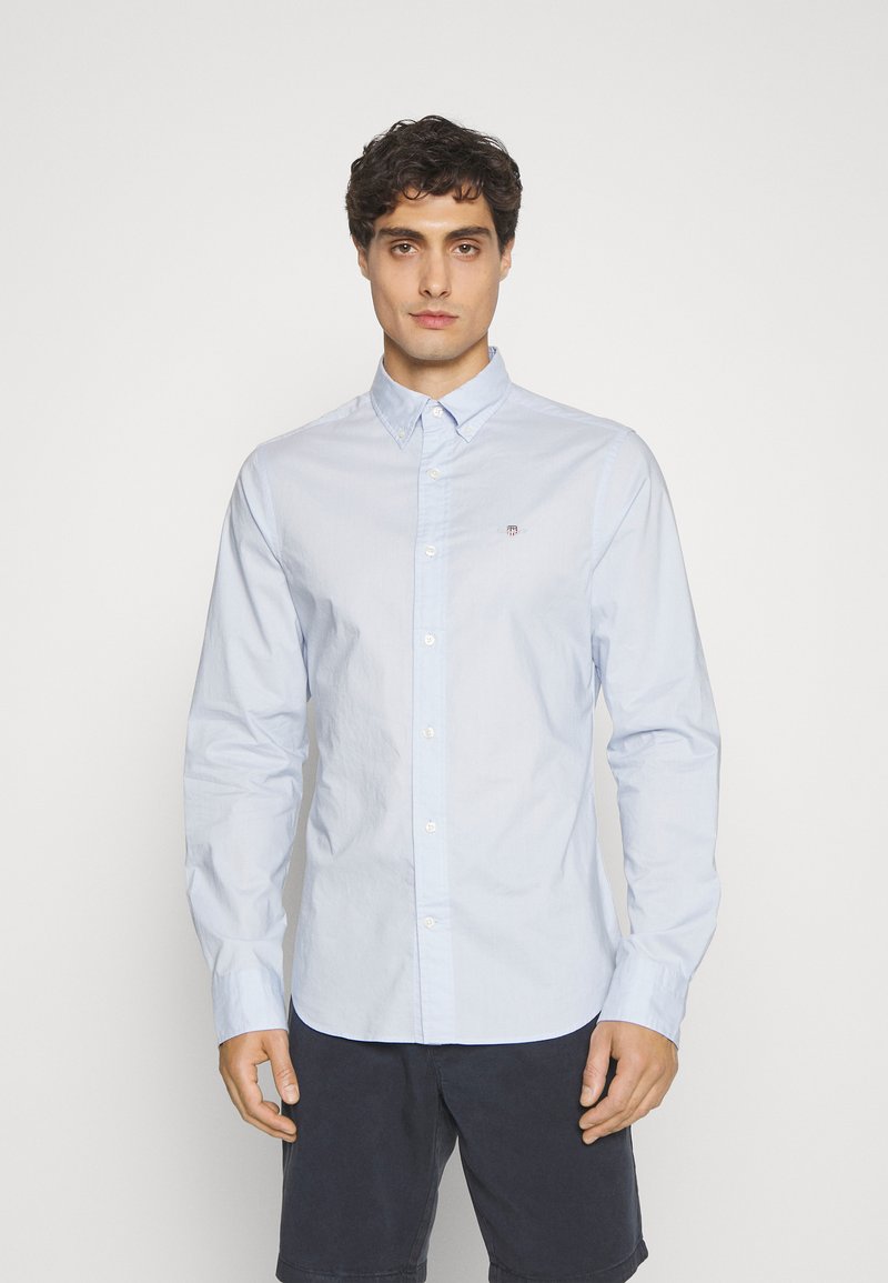 Jeune homme aux cheveux bouclés foncés, portant une chemise bleu clair boutonnée et un short foncé, debout devant un fond blanc uni.