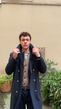 Joven con cabello rizado que ajusta el cuello de un abrigo azul marino de doble botonadura sobre un cárdigan beige, de pie en un patio con plantas.