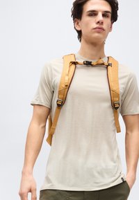 Νέος άντρας φορώντας μπεζ μπλουζάκι και πορτοκαλί σακίδιο πλάτης με λουρί στο στήθος, στέκεται μπροστά σε απλό ανοιχτό γκρι φόντο.