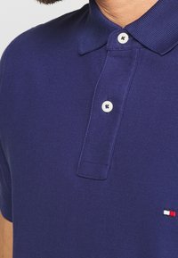 Polo shirt bleu marine en tissu texturé. Présente un col classique, une patte de boutonnage à deux boutons, et un petit logo brodé en rouge et blanc.