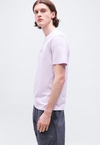 Νεαρός με μεσαίου μήκους καστανά μαλλιά φοράει ανοιχτό μωβ κοντομάνικο πουκάμισο και σκοτεινό γκρι παντελόνι, στέκεται μπροστά σε απλό λευκό φόντο.