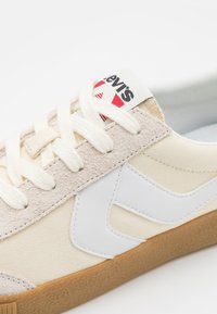 Zapatillas Levi's con parte superior de lona crema, acentos de ante beige, diseño de logo blanco y suela de goma texturizada con cordones blancos.