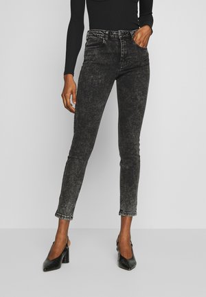 Osoba nosząca czarne obcisłe jeansy typu acid-wash, czarną bluzkę z długim rękawem oraz czarne czółenka na wysokim obcasie typu slingback, stojąca z jedną ręką w kieszeni.