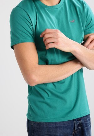 Zielona bawełniana koszulka z krótkim rękawem, okrągłym dekoltem i małym logo na klatce piersiowej. Noszona z ciemnoniebieskimi dżinsami, z założonymi rękami.