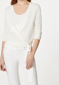 Top blanco con mangas de tres cuartos y detalle de lazo en la cintura, con una tela texturizada y diseño en capas sobre una camiseta de canalé.