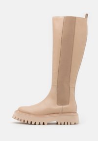 Beige polvisohva, jossa on paksu ura-ala ja leveä elastinen sivupaneeli, joka helpottaa pukemista, asetettuna yksinkertaista valkoista taustaa vasten.