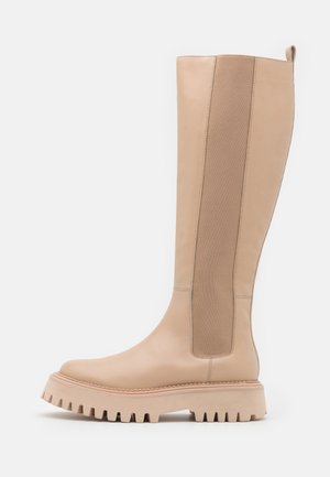 Beige kniehoog laars met stevige profielzool en brede elastische zijpaneel voor eenvoudig aan- en uittrekken, tegen een plain witte achtergrond.