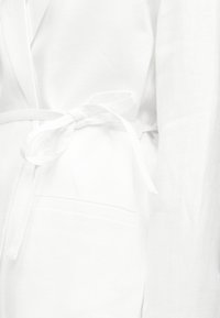 Gros plan sur une robe blanche nouée à la taille avec une ceinture en tissu par-dessus une manche longue.