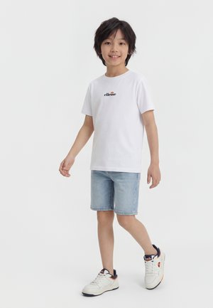 Băiat cu păr închis la culoare, zâmbind, purtând un tricou alb ellesse, pantaloni scurți din denim albastru deschis și adidași albi cu detalii portocalii și bleumarin.