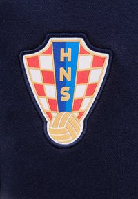Κεντημένο έμβλημα HNS με κόκκινο και άσπρο καρό μοτίβο, μπλε κάθετη γραμμή με τα γράμματα "HNS" και χρυσή πετοσφαιρική μπάλα στο κάτω μέρος.