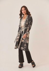 Mujer vistiendo un kimono negro con flores sobre una camiseta beige y pantalones negros, con sandalias de plataforma negras, de pie contra un fondo claro.