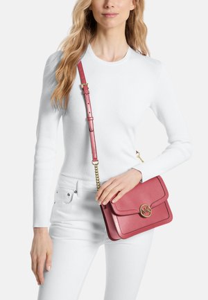 Mujer con top de manga larga blanco y pantalones, sosteniendo un bolso bandolera de cuero rosa con cadena dorada y cierre circular con logo.