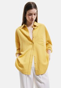 Gul button-up skjorte lavet af blødt stof, med krave, en brystlomme og opstegne ærmer. Sammen med hvide bukser.